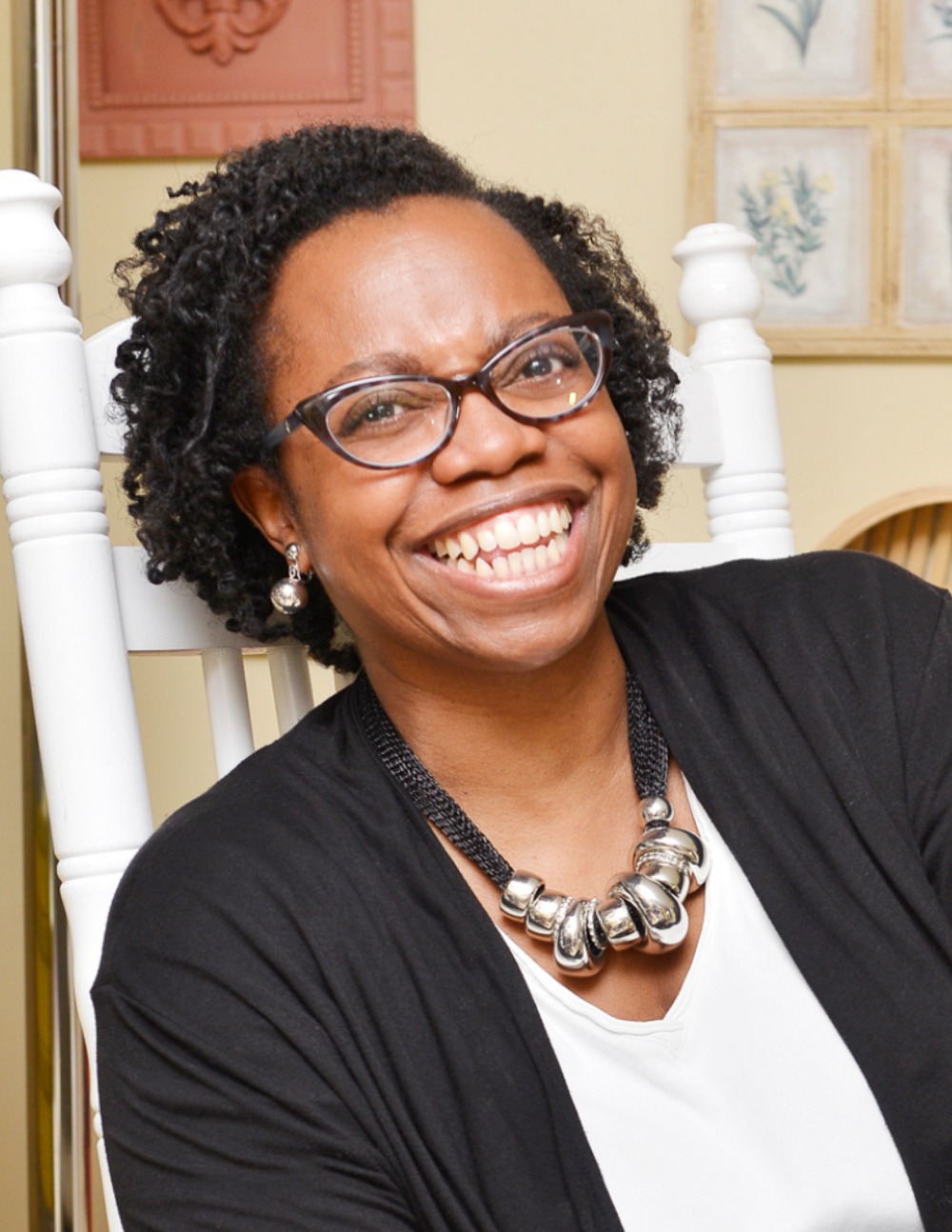 Smiling woman with glasses and short curly hair, wearing a chunky necklace and black cardigan, sits on a white chair in a warmly decorated room.
