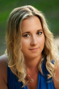 Portrait of a woman with wavy blonde hair, wearing a blue top, set against a blurred natural green background. Her expression is calm and serene.