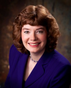 Smiling woman with wavy brown hair, wearing a purple blazer and necklace. The background is a textured, muted brown, creating a professional tone.