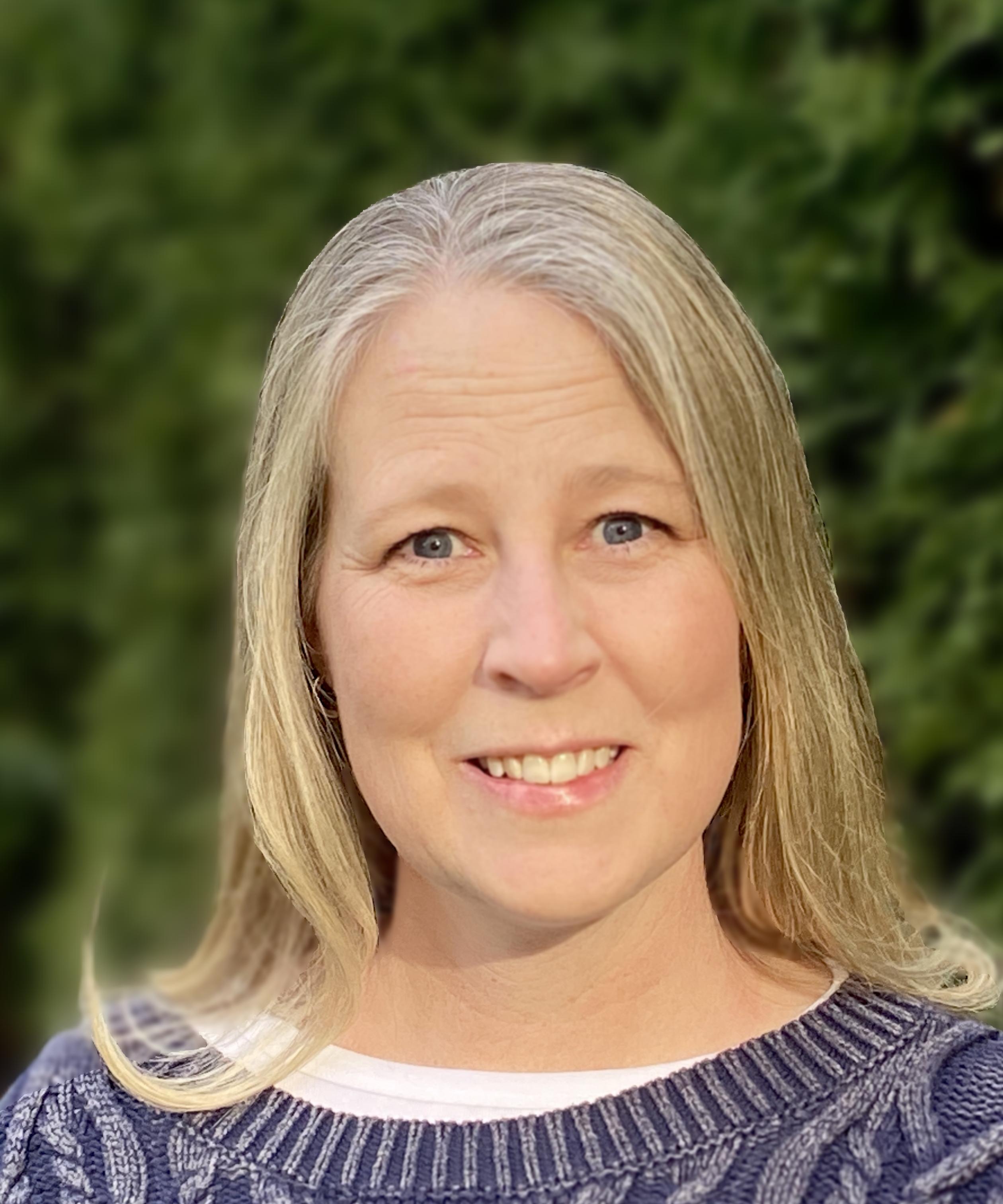 A woman with long blonde hair smiles subtly, standing outdoors against a lush green background. She wears a blue sweater, conveying a warm, friendly mood.