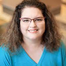 A woman with curly brown hair and glasses smiles warmly. She's wearing a blue top against a blurred neutral background, conveying a friendly, approachable vibe.