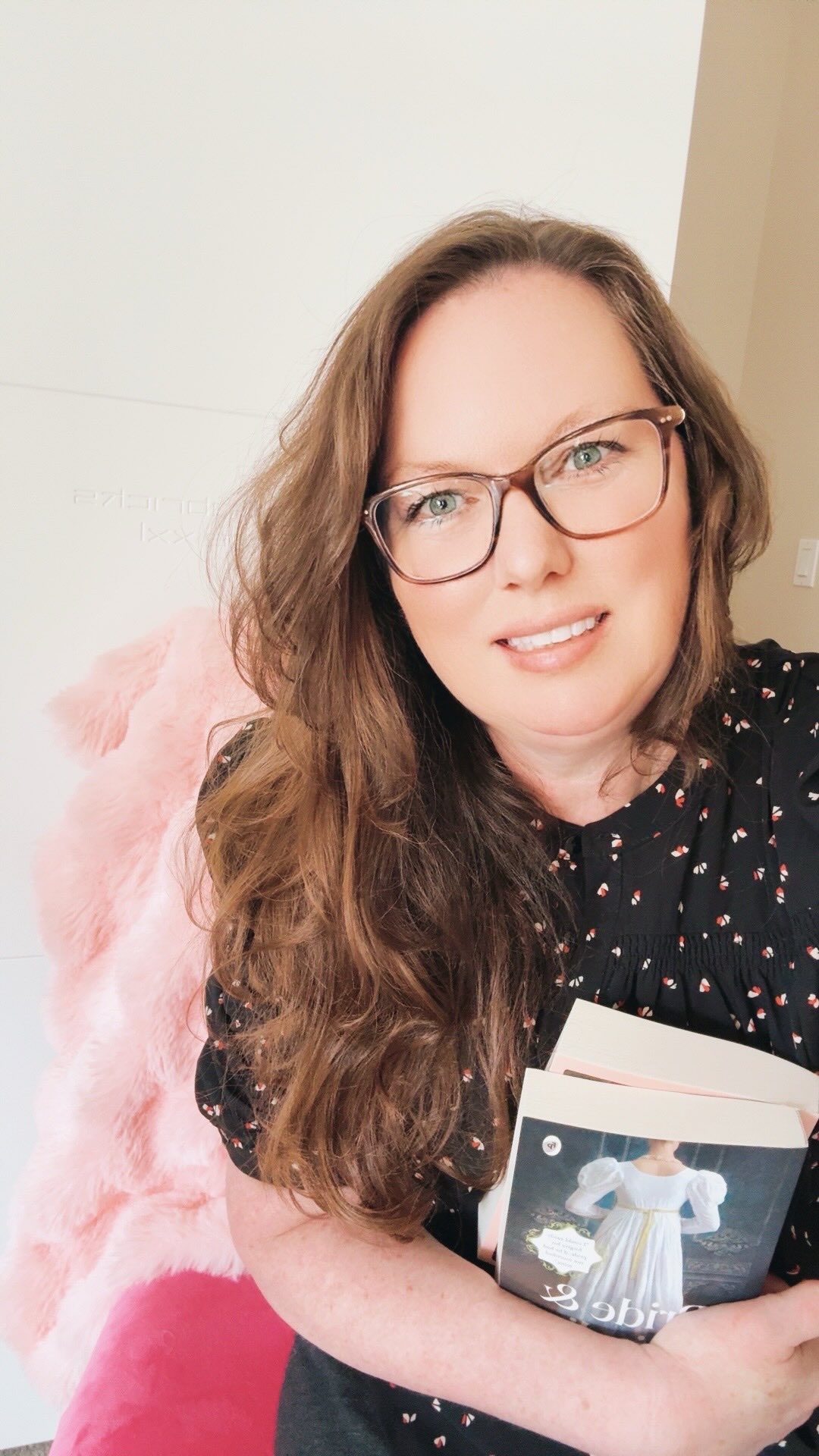 A woman with long brown hair and glasses smiles, holding a book. She wears a black top with small white designs. Behind her is a pink fluffy chair, creating a cozy atmosphere.