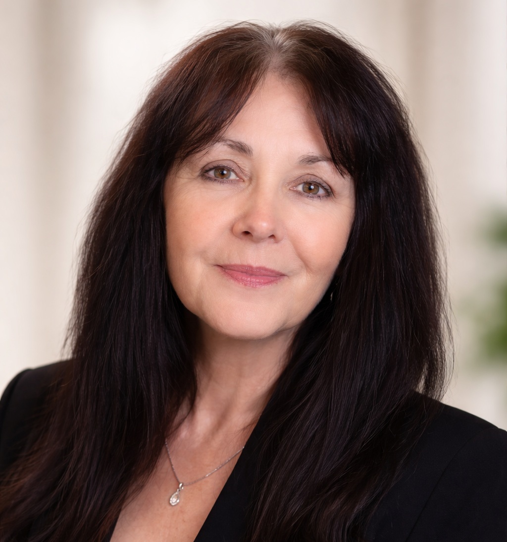 A woman with long dark hair and a gentle smile wears a black blazer and a delicate necklace. She stands against a softly blurred light background.