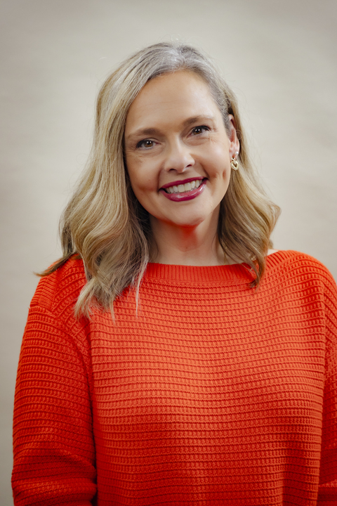 A woman with shoulder-length blonde hair smiles warmly at the camera, wearing a textured, bright orange sweater. The background is softly blurred, adding a gentle tone.