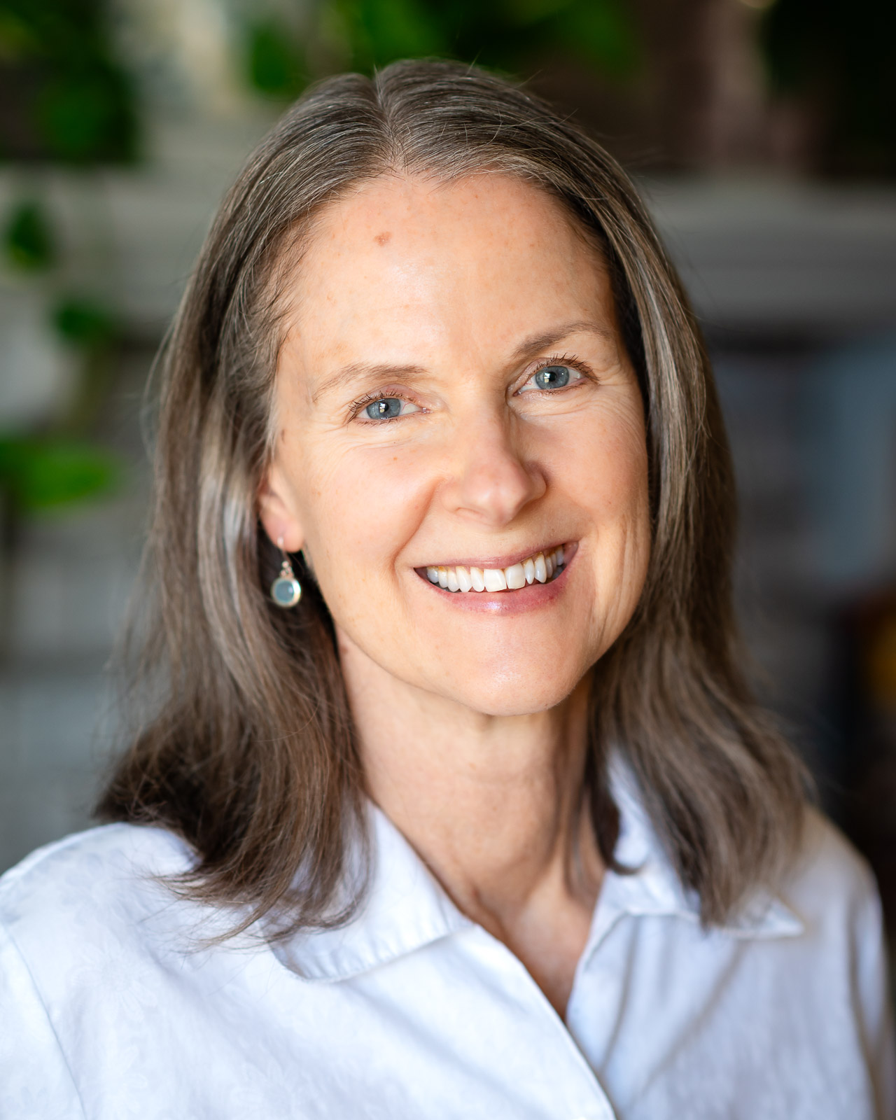 Smiling woman with long gray hair and blue eyes, wearing a white shirt and earrings. Soft lighting and a blurred green background convey warmth.