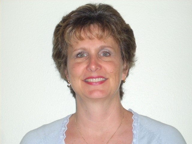 A woman with short brown hair smiling warmly at the camera, wearing a light blue top against a plain white background. The tone is friendly and welcoming.